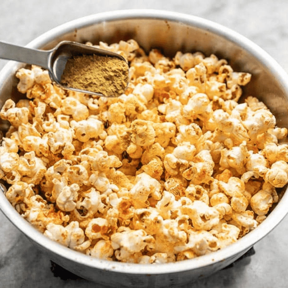 A bowl of popcorns with a spoon pouring nutritional yeast on it.